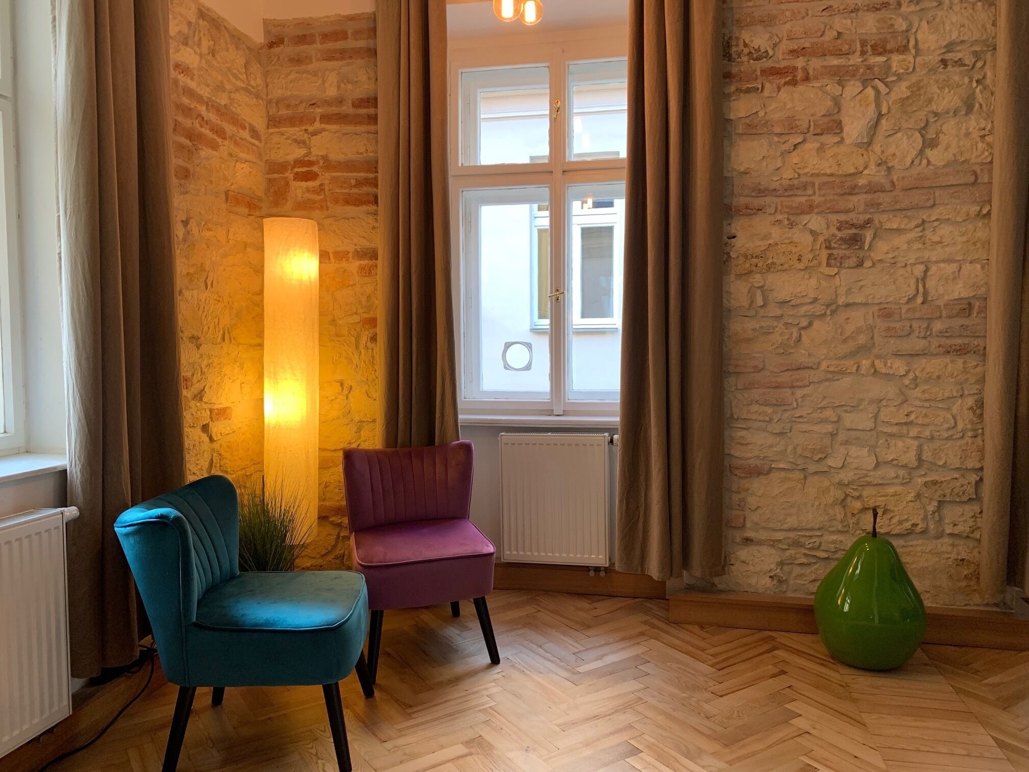 Ein Zimmer mit Fischgrätparkett, einem türkisfarbenen Sessel, einem violetten Sessel, einer grünen Vase und einer Ziegelwand.