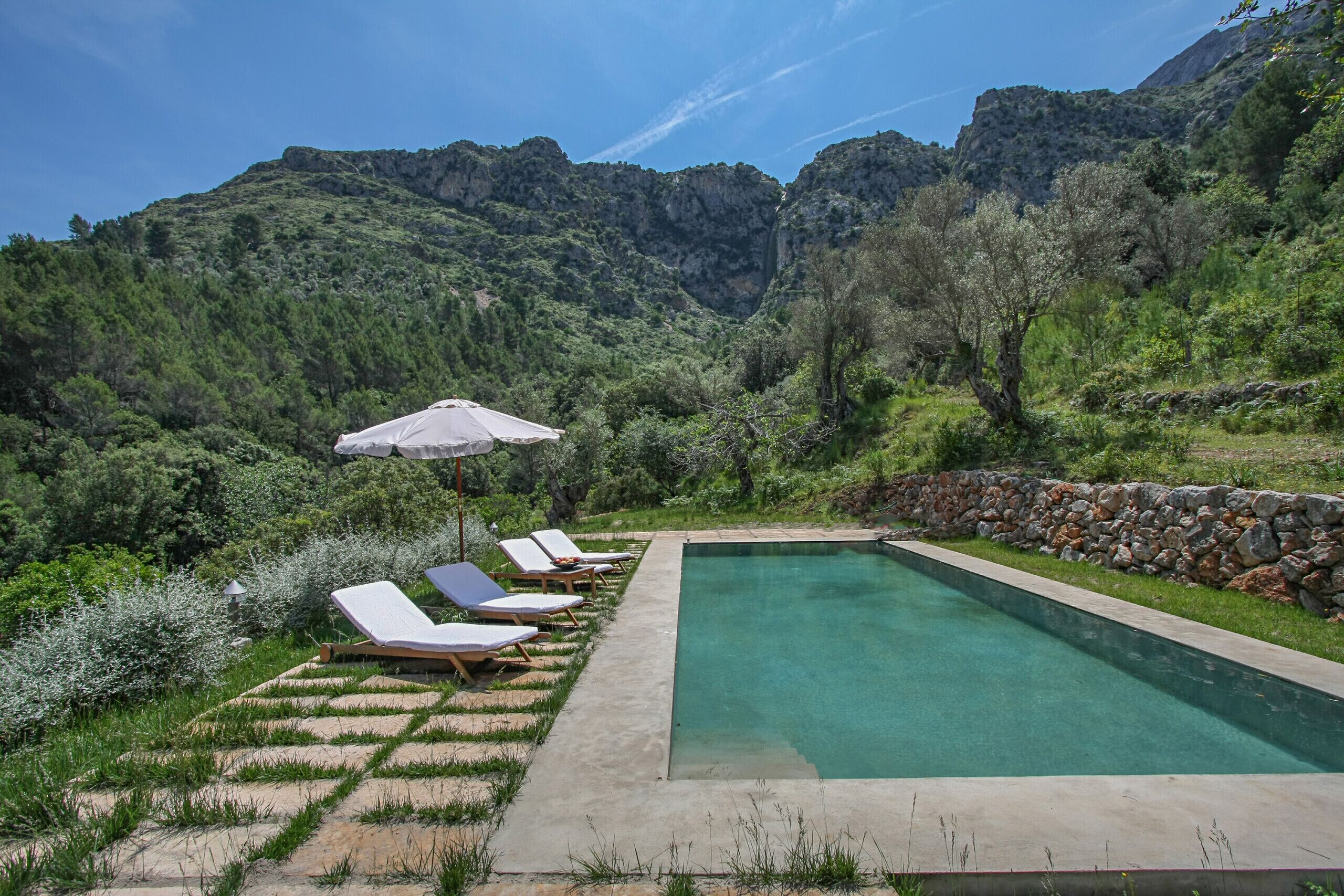 A rectangular pool with lounge chairs and an umbrella, surrounded by a stone wall and lush greenery, with mountains in the background.