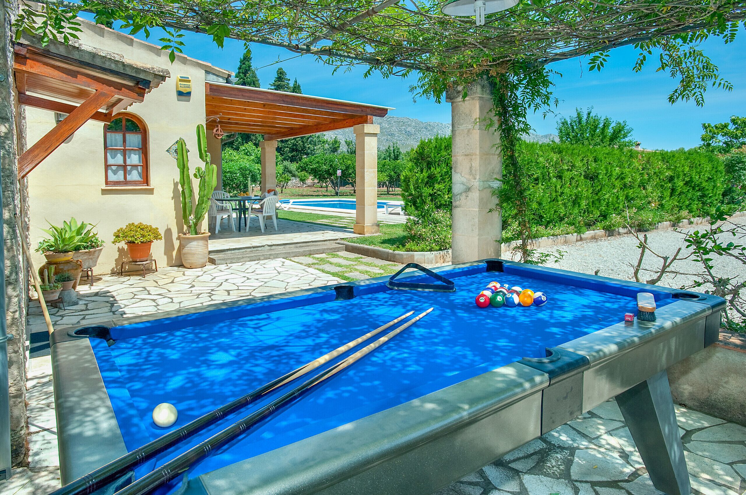A pool table with pool cues and balls, a patio with outdoor furniture, and a view of a pool and mountains in the background.
