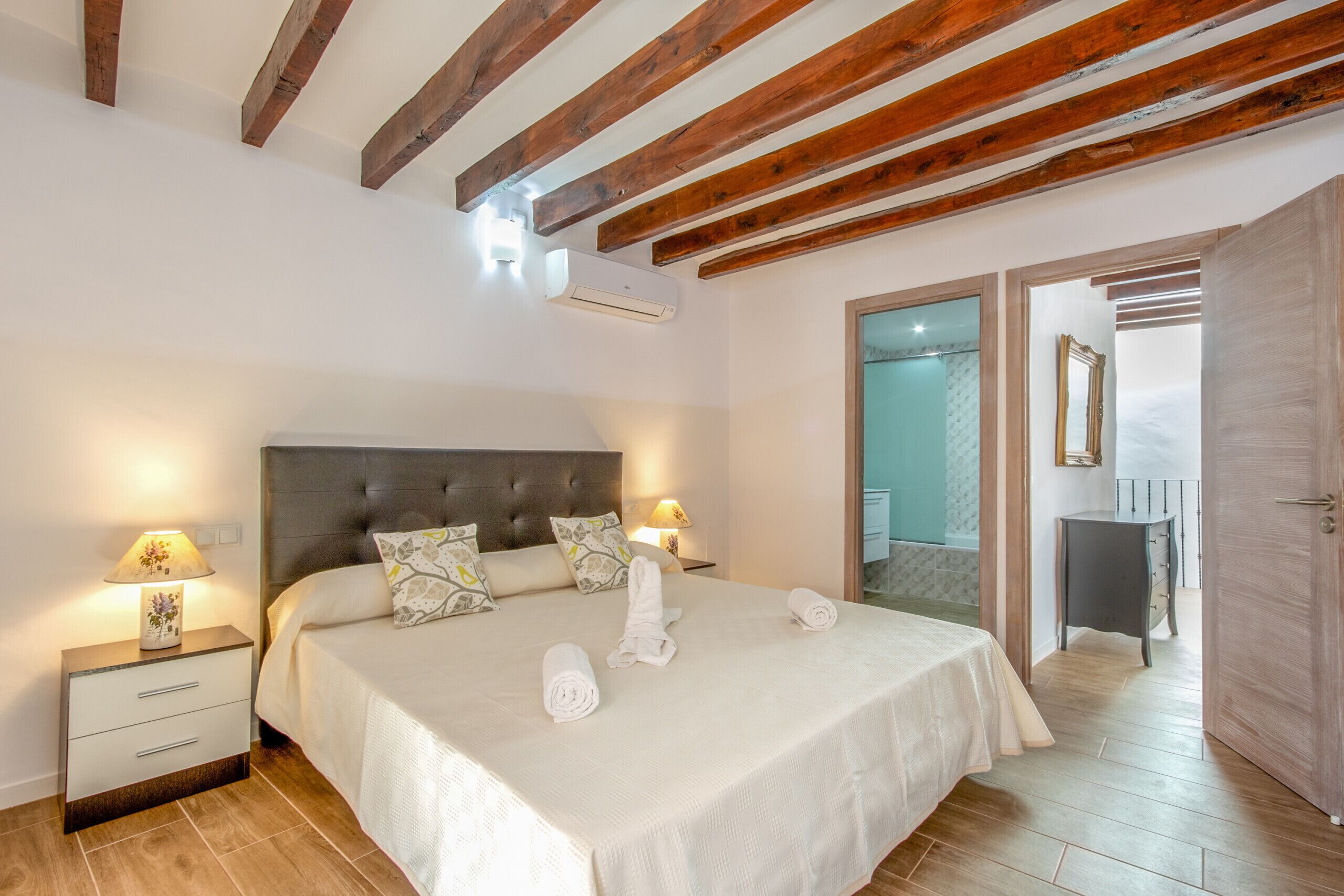 Una camera da letto con un letto grande, travi di legno a soffitto, due comodini con lampade, e un bagno visibile attraverso una porta aperta.