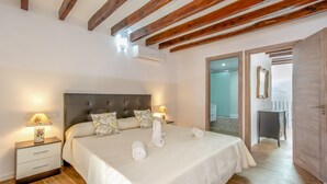 A bedroom with a large bed, wooden beams on the ceiling, two bedside tables with lamps, and a bathroom visible through an open door. - Town55, House 5StarsHome Mallorca (Pollensa / Pollença)