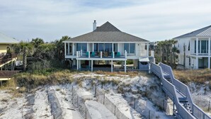 Uma casa de praia de dois andares com varanda, rodeada por dunas de areia e vegetação.