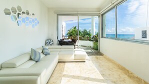 A modern living room with a white sofa, a ceiling fan, and a large window offering a view of palm trees and the ocean. - Beachfront Condo w/ Large Balcony and Ocean Views near Downtown Playa Del Carmen (Playa del Carmen)