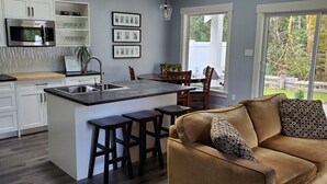 A modern kitchen with white cabinets, a built-in island with stools, and a dining area with a brown couch and patterned pillows. - A modern cozy cottage in your own wooded backyard located mid Vancouver Island. (Qualicum Beach)