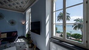 Ein Zimmer mit großem Fenster mit Blick auf ein Gewässer, einer Decke mit dekorativen Paneelen, einem schwarzen Sofa und einem an der Wand befestigten Fernseher.