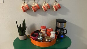Una zona de cocina con una mesa verde, un colgador para tazas, una cafetera y diversos utensilios de cocina.