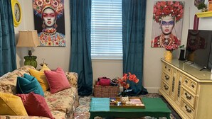 Una sala de estar con un sofá floral, una mesa de centro verde, un tapete estampado, un televisor sobre un mueble de madera y obras de arte en las paredes.