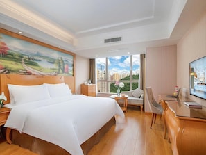 A hotel room with a large bed, a desk, a chair, a TV, and a window with a city view.