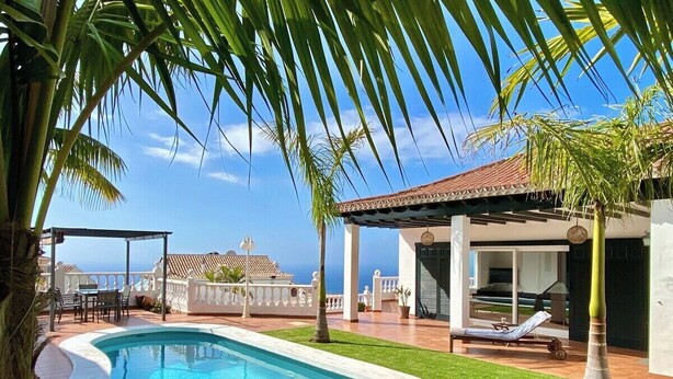 A swimming pool with a white edge, surrounded by a well-maintained lawn and palm trees. There is a covered patio area with outdoor furniture and a view of the sea. - Home2Book Design Asian Sabina, Heated Pool & View (El Sauzal)