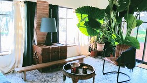 Un salon chaleureux avec un banc en bois, une petite table avec des boissons, une lampe et de grandes plantes en pot.