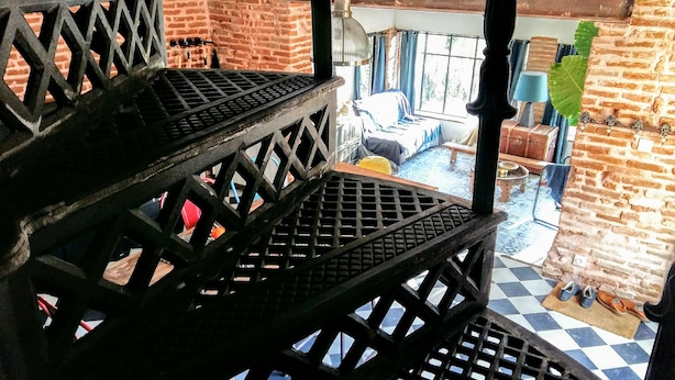 Un escalier avec une rampe en métal mène à une pièce dont le sol est à carreaux, un mur est en briques et une fenêtre est ornée de rideaux.