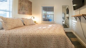 A bedroom with a bed, a bedside table, a mirror, a TV mounted on the wall, and a window with blinds. - Home in the Historic Coronado District 2bed,1 bath (Phoenix)