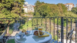 Un balcón con una mesa dispuesta para el desayuno, incluyendo croissants y un vaso de zumo de naranja, con vistas a un jardín y edificios.