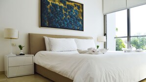 A neatly made bed with white linens, a beige headboard, and a large abstract painting above it. There are two bedside tables with lamps and a small plant on one. - Menesse 25.26 (Playa del Carmen)