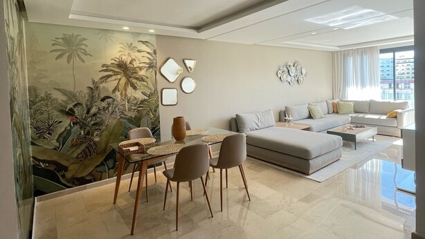 A modern living room with a dining area, a large sofa, and a mural of tropical plants. - Cozy and beautiful apartment 2 steps from la corniche (Dar-el-Beida)