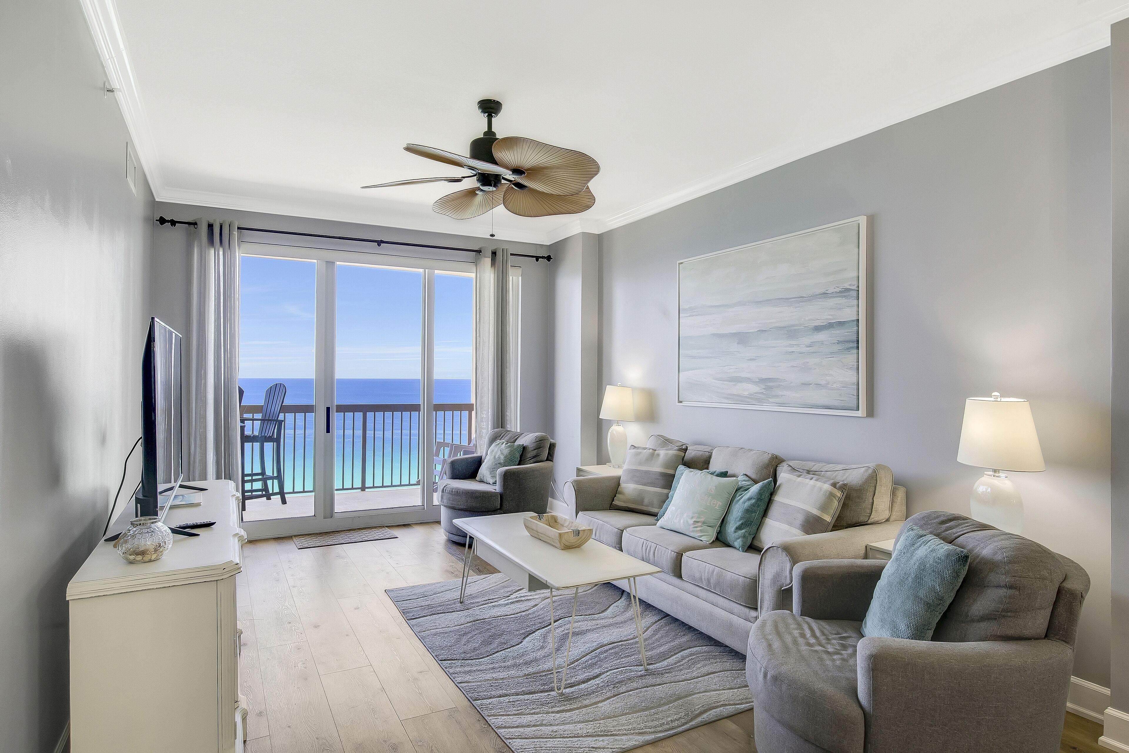 A modern living room with a balcony, a ceiling fan, and a large window offering a view of the ocean.