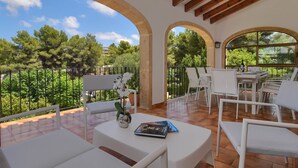 Une terrasse extérieure avec des meubles blancs, une table sur laquelle se trouvent un livre et un vase, et une vue sur la verdure et les arbres.