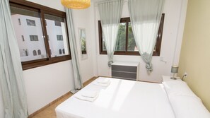 Habitación de hotel con cama, mesita de noche con lámpara, ventana con cortinas y vistas a edificios.