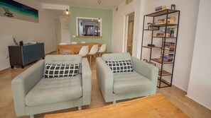 A living room with two armchairs, a wooden coffee table, a dining table with chairs, a bookshelf, and a mirror.