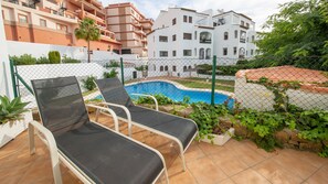 Un área junto a la piscina con dos sillas de descanso y una pileta, rodeada por una cerca de alambre y follaje.