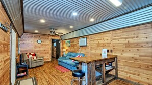 A cozy cabin interior with a wooden floor, a blue sofa, a wooden table with stools, a ceiling fan, and a clock on the wall.