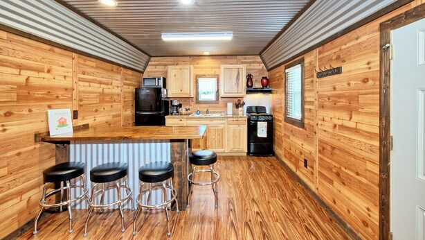 A compact kitchen with wooden walls, a central island with stools, a refrigerator, a microwave, a stove, and a sink.