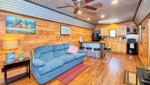 A cozy cabin interior with a blue sofa, wooden walls, a ceiling fan, a kitchen area, and a painting of a barn.