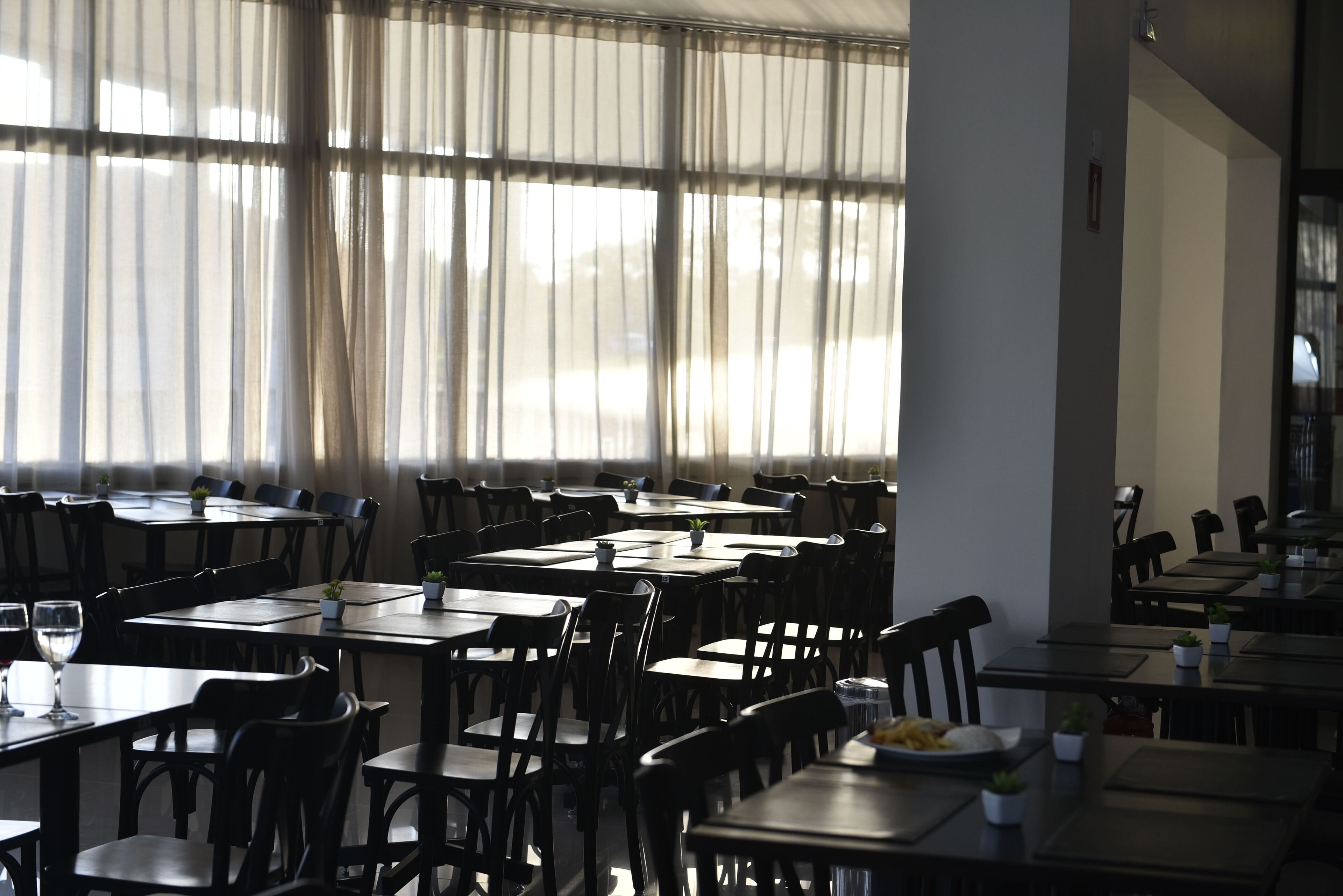 A restaurant with multiple tables and chairs, large windows with sheer curtains, and potted plants on the tables.