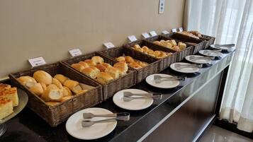 A buffet with various breads and pastries in wicker baskets, arranged on a counter with plates and tongs.