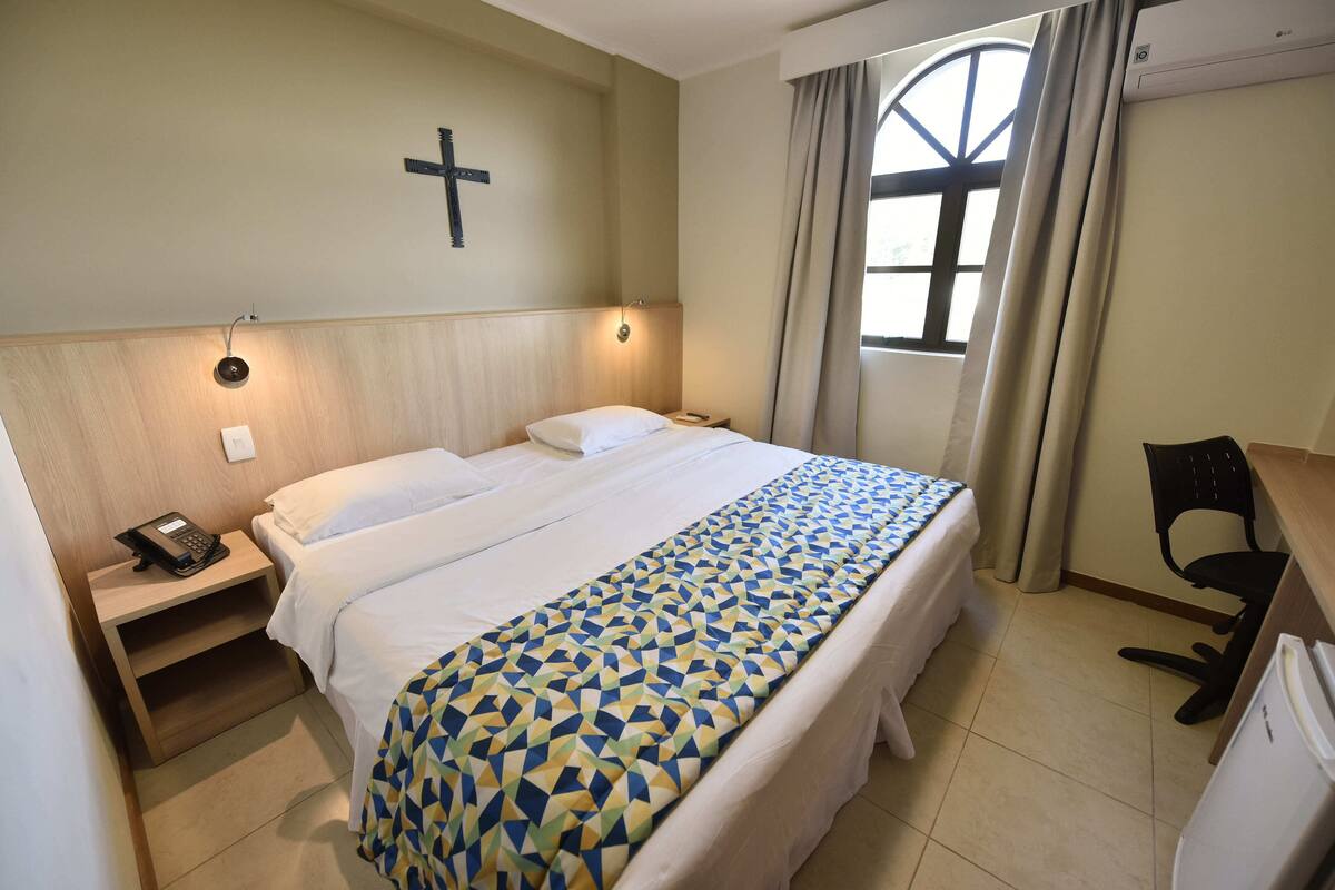 A hotel room with a bed, a desk, a chair, a phone, and a cross on the wall.