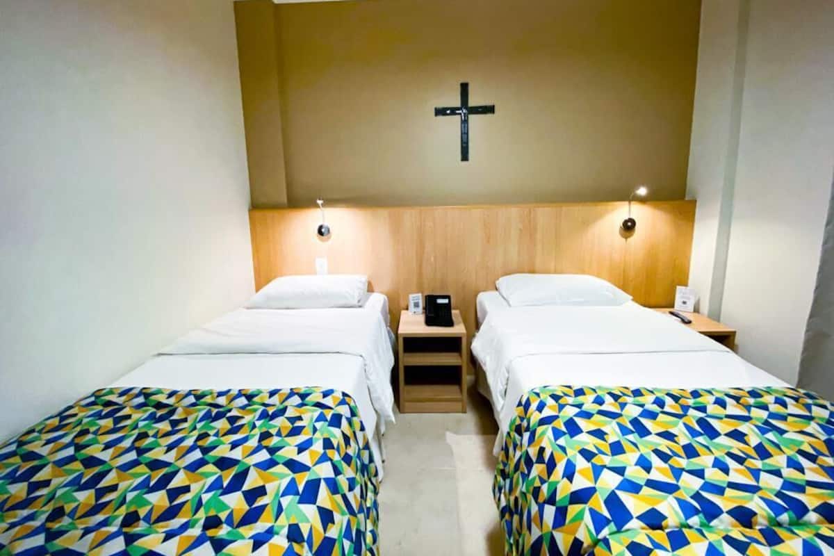 A hotel room with two beds, a wooden headboard, a cross on the wall, and bedside tables with lamps.
