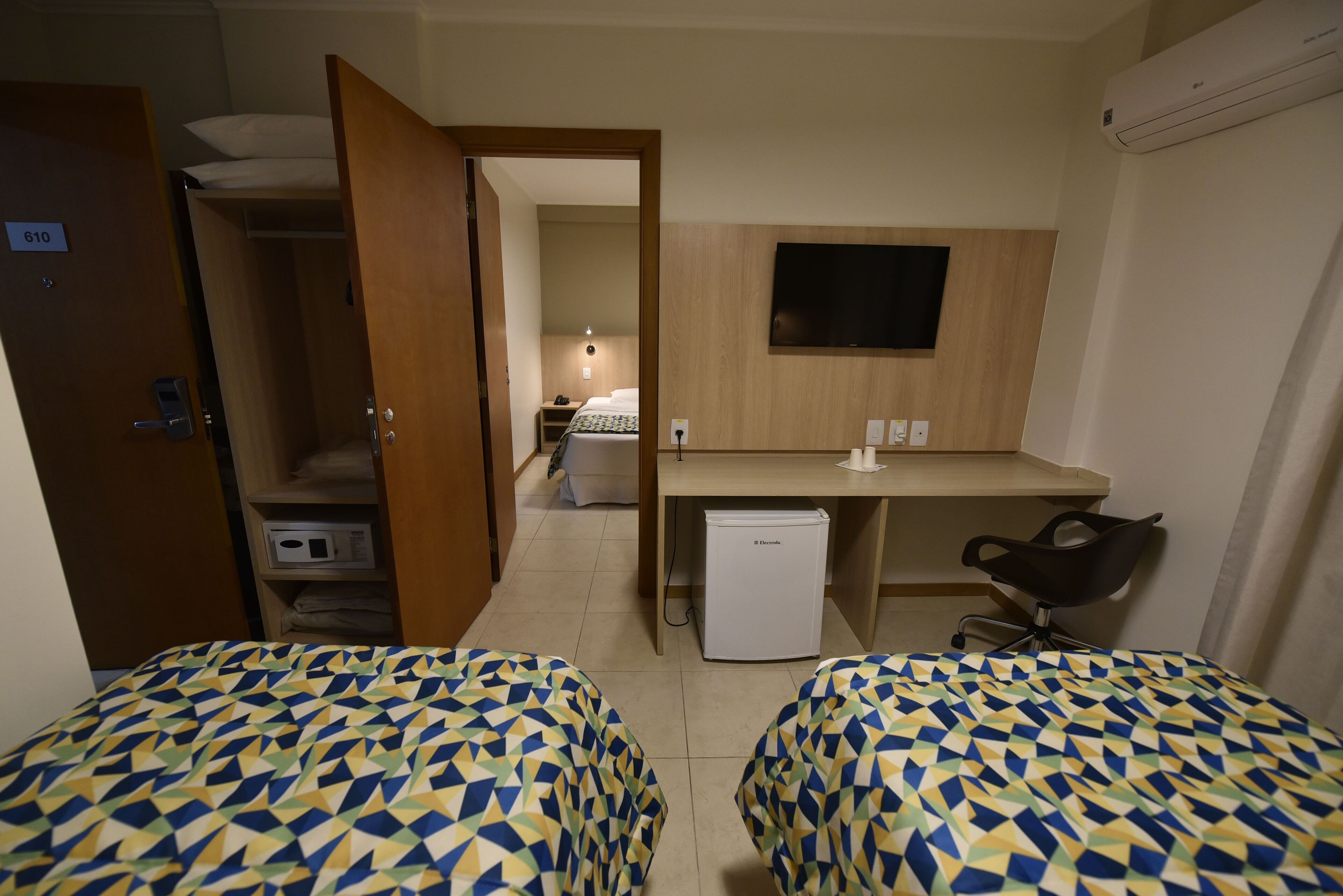 A hotel room with two beds, a desk, a chair, a refrigerator, and a TV.