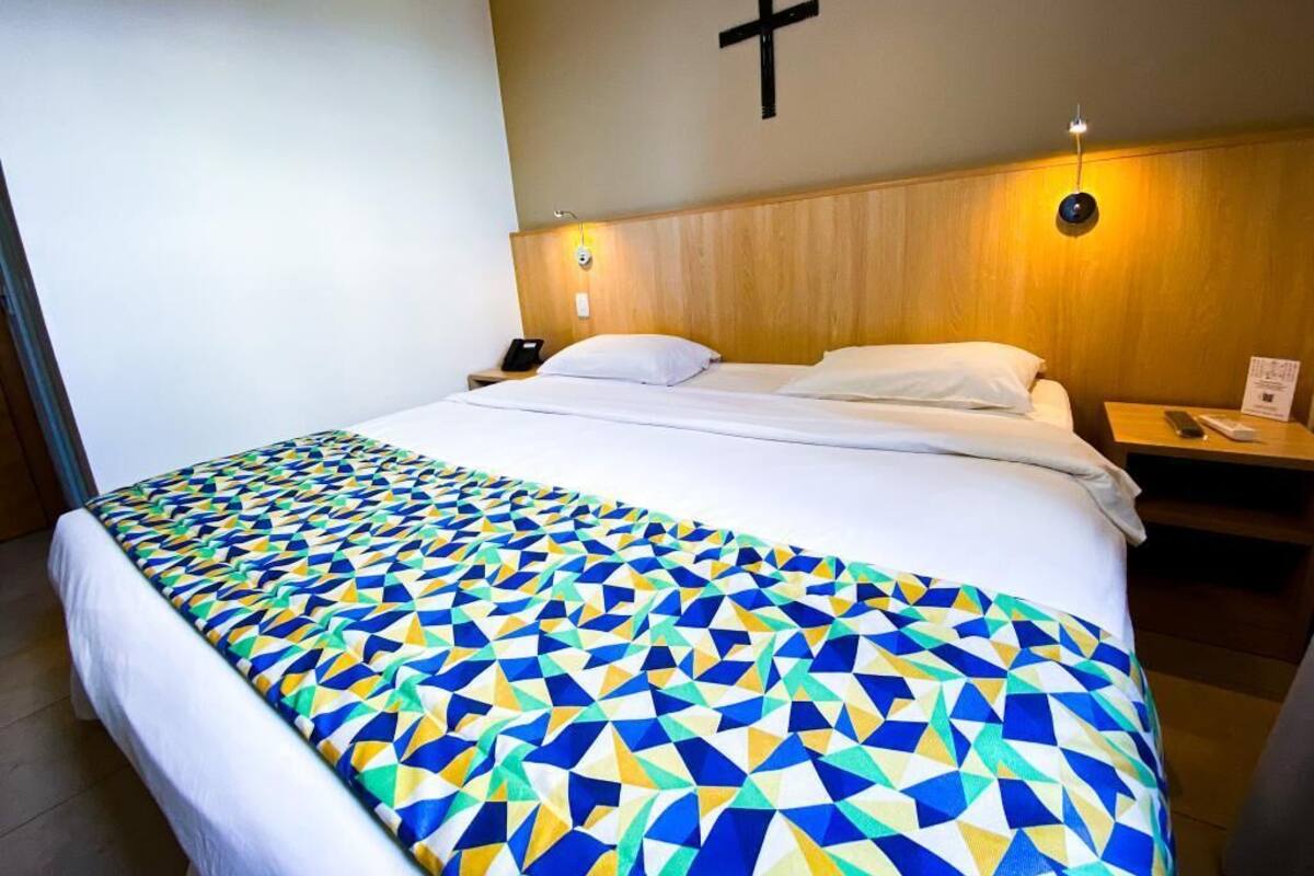 A hotel room with a bed, a wooden headboard, a cross on the wall, and a nightstand with a lamp.