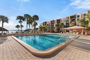 A swimming pool with a tiled deck, surrounded by palm trees and a building with balconies.