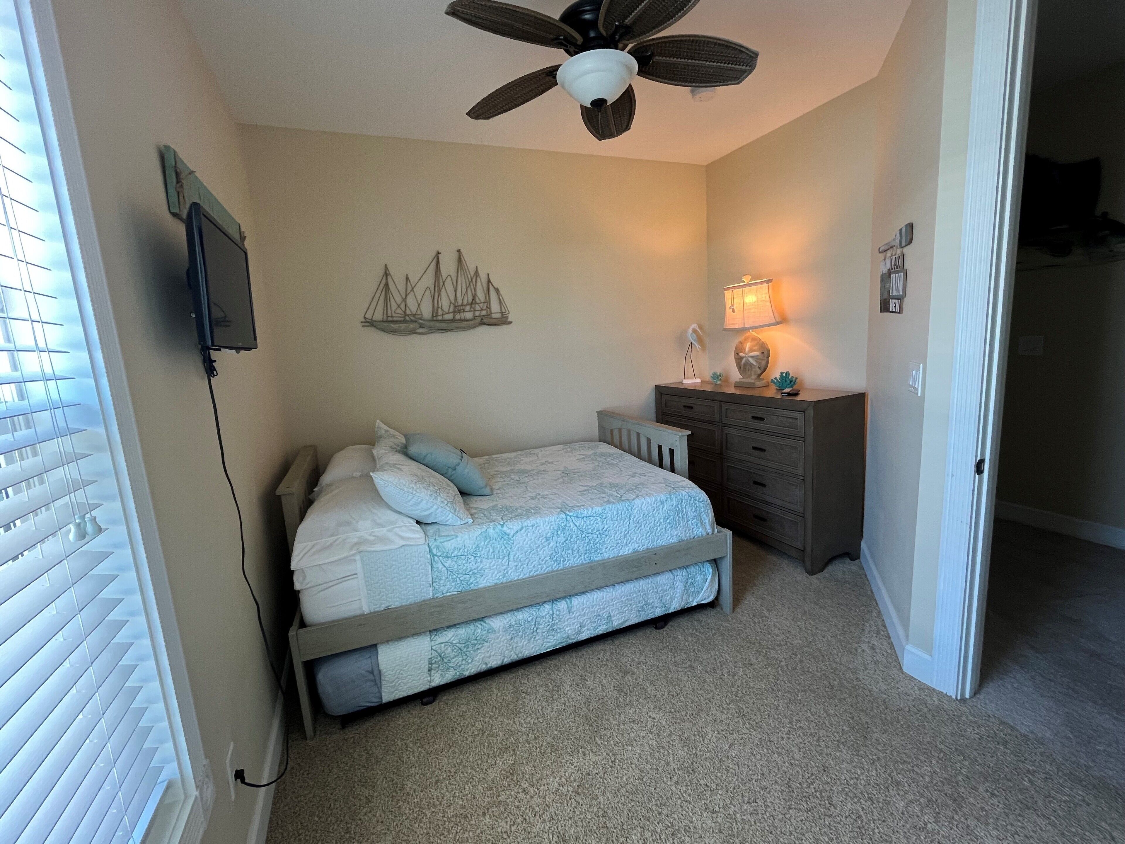 A bedroom with a bed, a dresser, a ceiling fan, a wall-mounted TV, and a window with blinds.