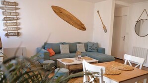 A cozy living room with a sofa, coffee table, and dining table. There are decorative items like a surfboard, paddle, and a sign with beach-related words. - Le 8 ∙ Cosy apartment with sea-view balcony (Mers-les-Bains)