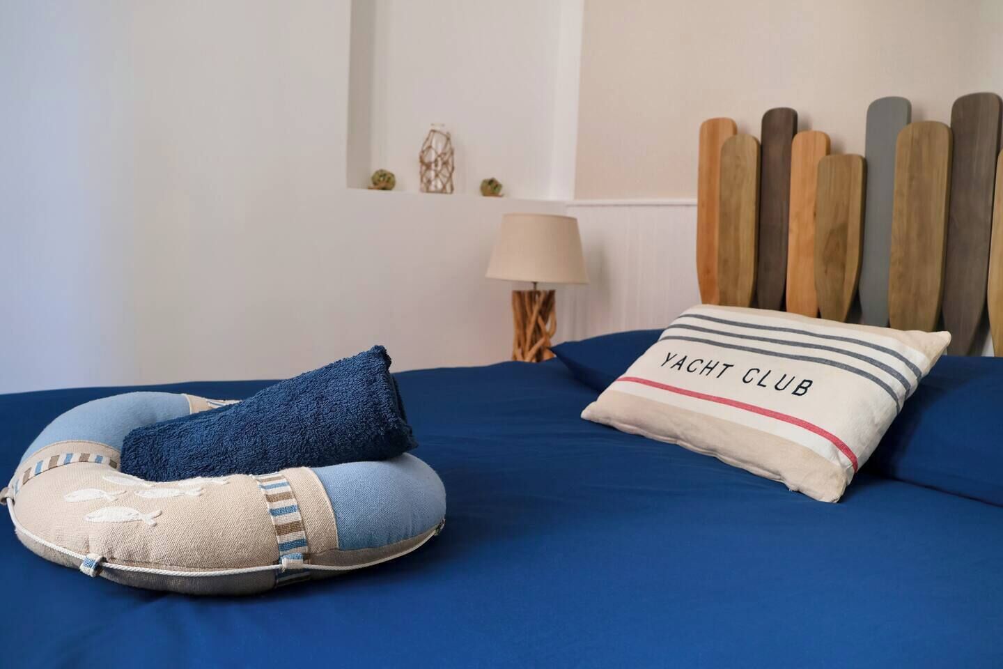 Een bed met een blauwe spreideken, een kussen met de tekst 'YACHT CLUB' en een hoofdbord met houten latten.