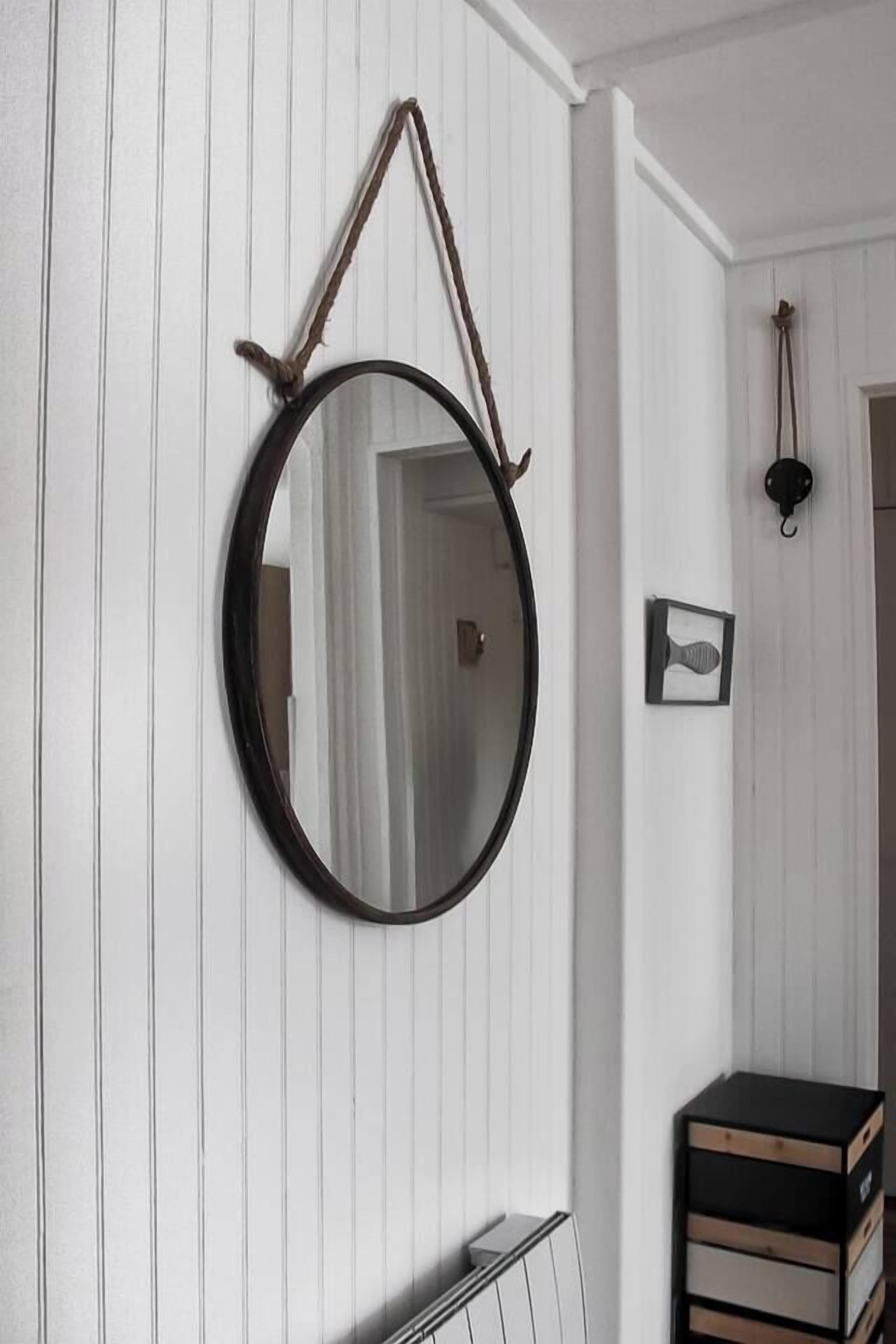 Un miroir rond avec une anse en corde, fixé sur un mur blanc à panneaux.