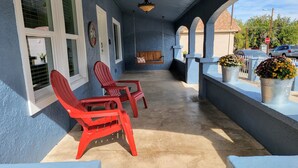 Eine überdachte Veranda mit zwei roten Adirondack-Stühlen, einer hölzernen Bank und einem Metallkübel mit Blumen.