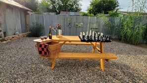 Una mesa de picnic de madera con piezas de ajedrez y una botella de vino, sobre grava con una valla de metal corrugado al fondo.