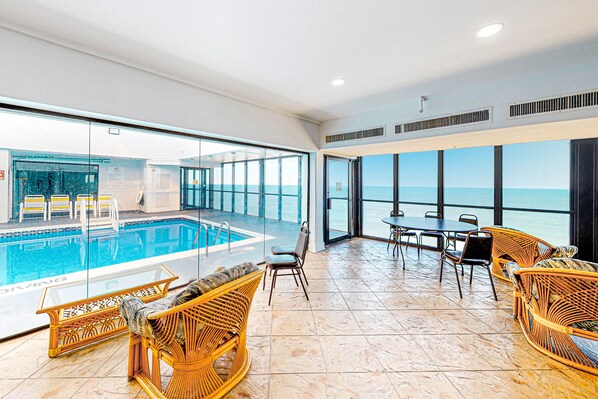 Relax in a spacious area with a stunning ocean view through a large glass wall, featuring a pool and comfortable wicker furniture.