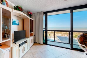 Relax in this stylish living area with a view of the beach and pool, featuring a flat-screen TV, comfortable chair, and dining table for two.