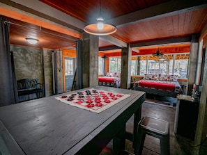 Une table à manger en bois, agrémentée d’un chemin de table à carreaux et d’un jeu d’échecs. On aperçoit en arrière-plan deux lits avec une literie rouge et blanche.