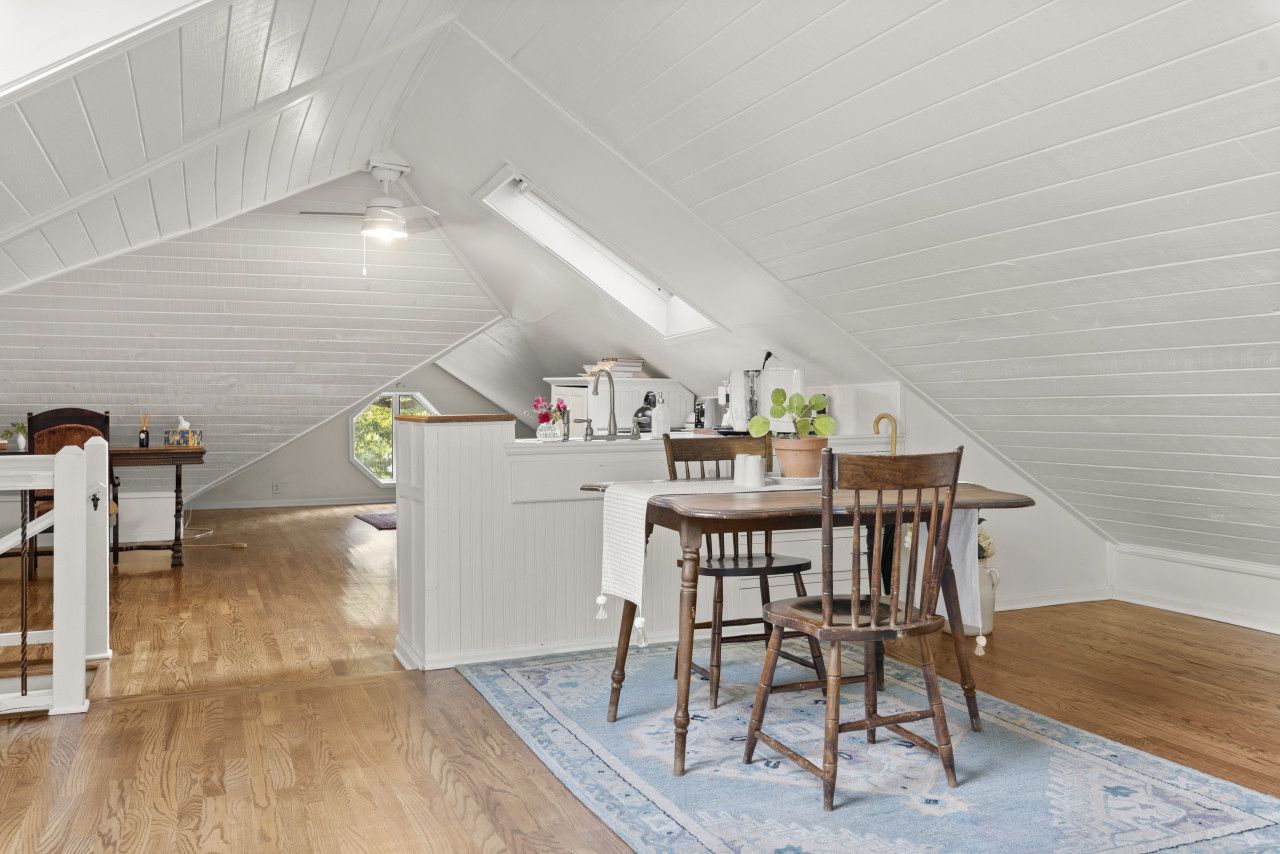 An attic bedroom with a sloped ceiling, wooden flooring, a dining table with chairs, a white dresser, and a window.