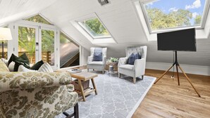 A cozy attic room with a high ceiling, skylights, a comfortable sofa, two armchairs, a small table, and a flat-screen TV on a tripod stand.