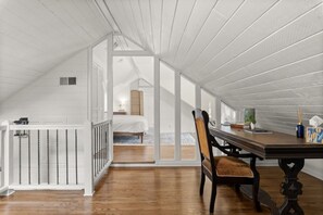 A room with a wooden floor, a dining table with chairs, a white railing, and a bed in the background.