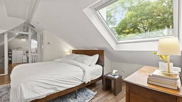 A bedroom with a skylight, a bed, a bedside table with books, and a view of trees outside.
