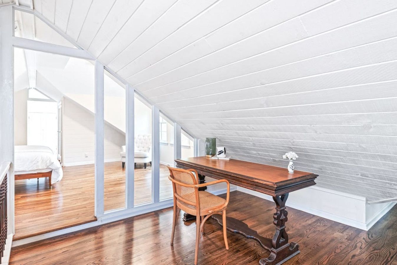 A spacious attic bedroom with a wooden floor, a large window, a bed, a desk, and a chair.