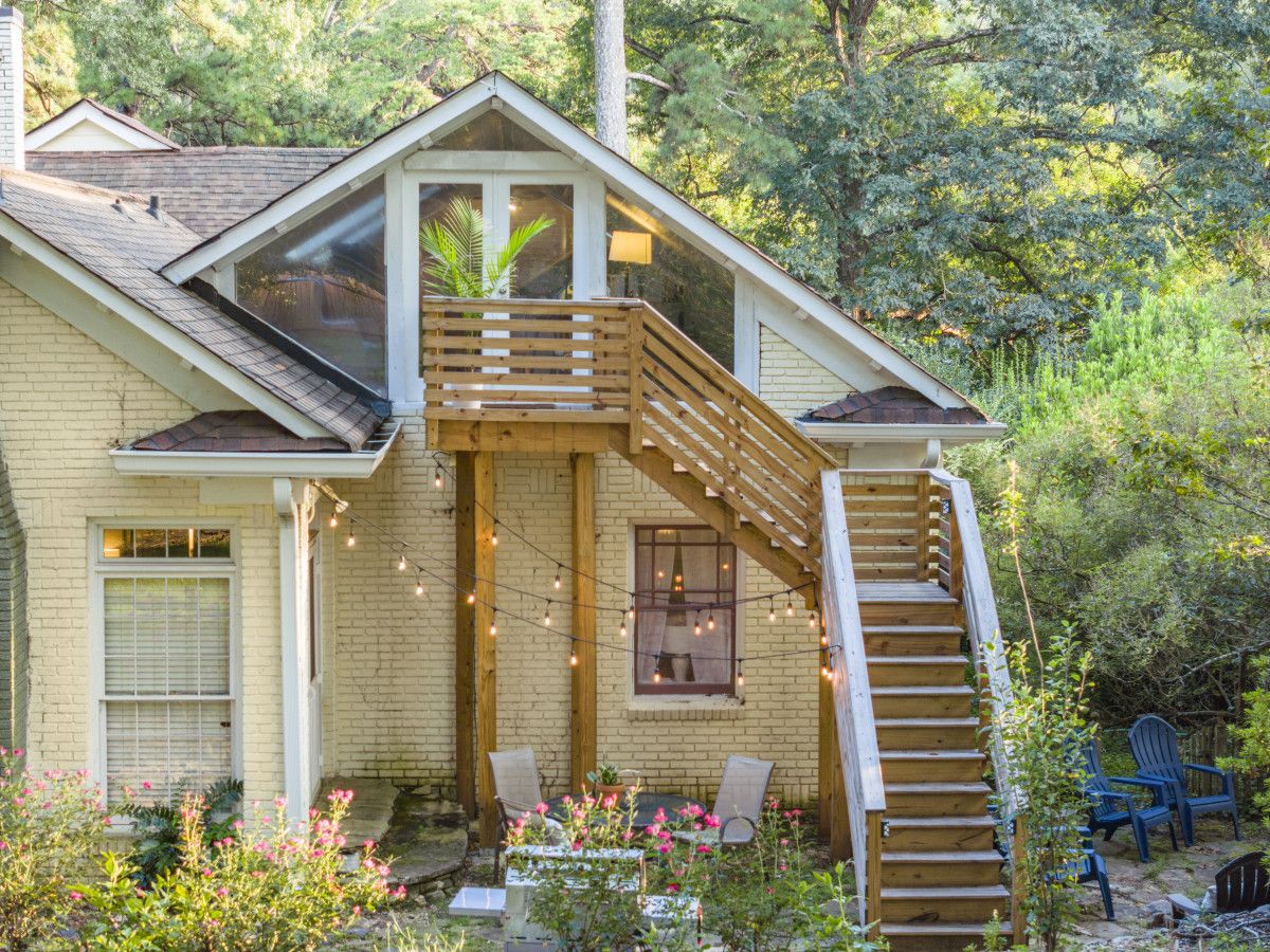 A two-story house with a wooden staircase leading to a balcony, surrounded by greenery.