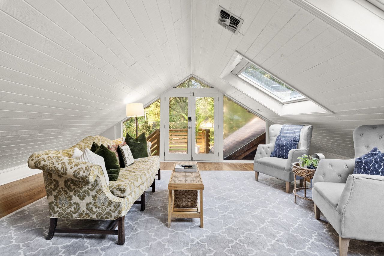 Une chambre mansardée avec un plafond en pente, une grande porte-fenêtre coulissante donnant sur une terrasse, un canapé à motifs, un fauteuil blanc et une petite table.